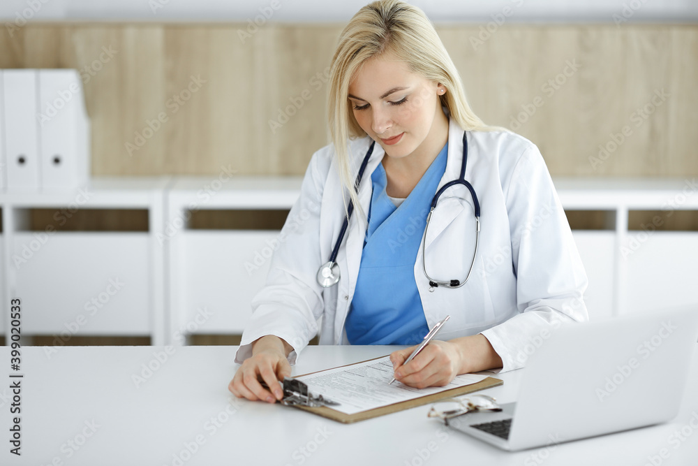 Portrate of woman-doctor at work while sitting at the desk in clinic. Blonde cheerful physician ready to help patients Portrate of woman-doctor at work while sitting at the desk in clinic. Blonde cheerful physician ready to help patients
