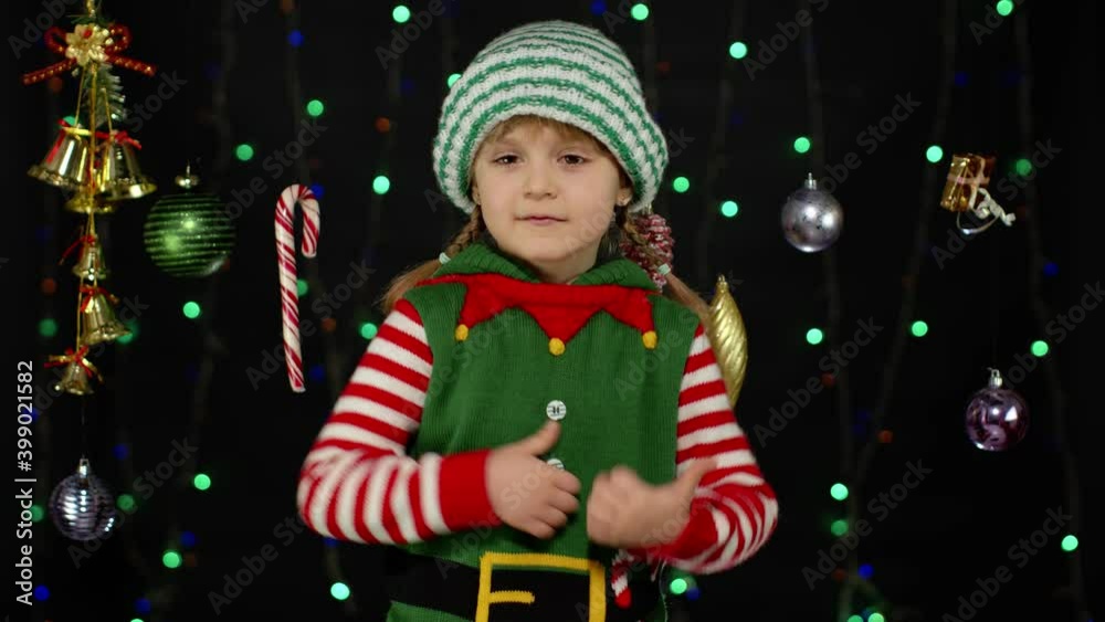 Teen kid girl in Christmas elf Santa Claus helper costume showing thumbs up gesture, agree sign isolated on black background with garland decorations toys. Child smiling. New Year holidays celebration Teen kid girl in Christmas elf Santa Claus helper costume showing thumbs up gesture, agree sign isolated on black background with garland decorations toys. Child smiling. New Year holidays celebration
