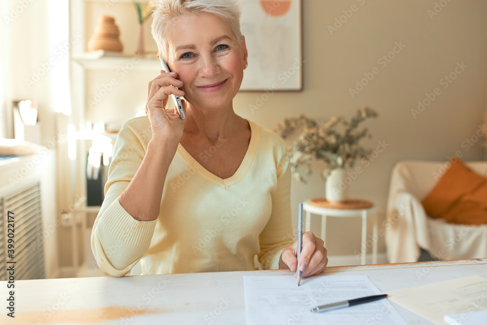 Attractive middle aged female manager sitting at table with documents, looking at camera with pleased smile, holding mobile, making phone call, talking to supplier while working on commercial offer