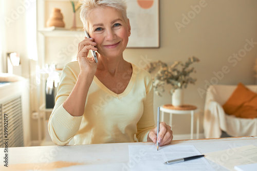 Attractive middle aged female manager sitting at table with documents  looking at camera with pleased smile  holding mobile  making phone call  talking to supplier while working on commercial offer