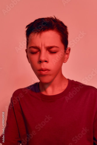 Close up portrait of teenaged disabled boy with cerebral palsy keeping his eyes closed, suffering from pain, posing isolated over red light background