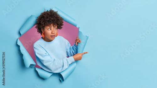 Surprised dark skinned Afro American woman looks with great wonder and advertises item shows copy space for your promotion dressed in long sleeved jumper poses through paper hole. Breakthrough