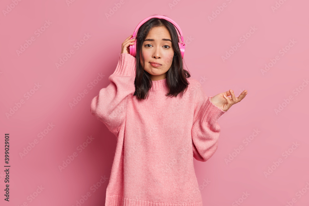 Confused dark haired woman raises palm and feels hesitant cannot remember whether she listened this song before or not wears stereo headphones dressed casually isolated over pink background. Confused dark haired woman raises palm and feels hesitant cannot remember whether she listened this song before or not wears stereo headphones dressed casually isolated over pink background.