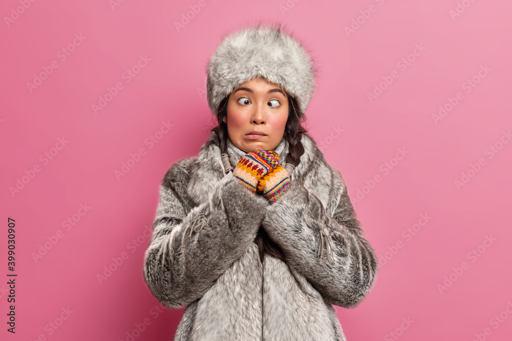 Funny crazy Eskimo woman makes grimace crosses eyes wears winter fur clothes has fun in studio poses against pink background. Arctic female plays fool dressed in coat and hat for frosty weather Funny crazy Eskimo woman makes grimace crosses eyes wears winter fur clothes has fun in studio poses against pink background. Arctic female plays fool dressed in coat and hat for frosty weather
