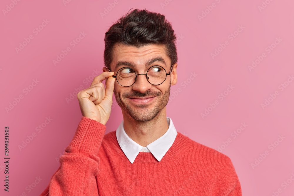Φωτογραφία Headshot of handsome European man with bristle looks aside and smiles pleasantly has thoughtful expression wears round spectacles red jumper isolated over pink background