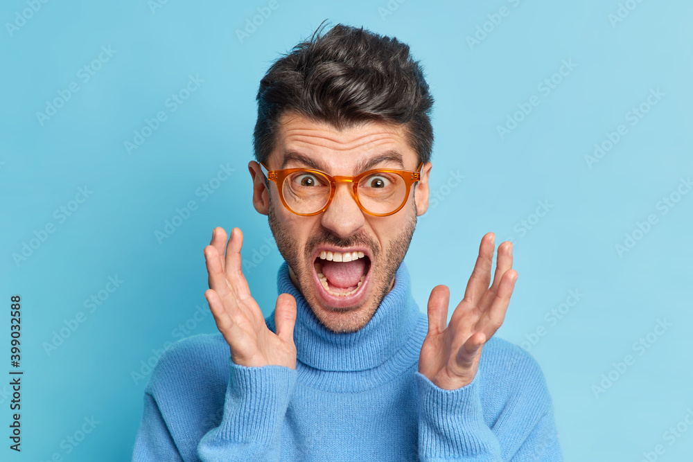 Φωτογραφία Close up portrait of displeased young man raises hands and screams loudly being annoyed with something keeps mouth widely opened wears optical glasses and turtleneck poses indoor