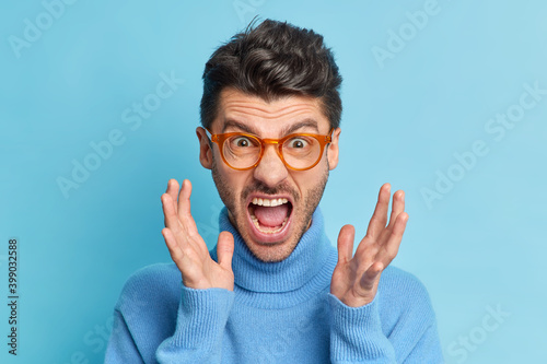 Fotótapéta Close up portrait of displeased young man raises hands and screams loudly being annoyed with something keeps mouth widely opened wears optical glasses and turtleneck poses indoor