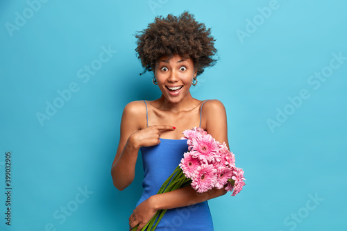 Is it for me. Amazed cheerful African American woman points at herself cannot believe in receiving positive news holds beautiful pink gerbera flowers wears blue dress poses with bare shoulders