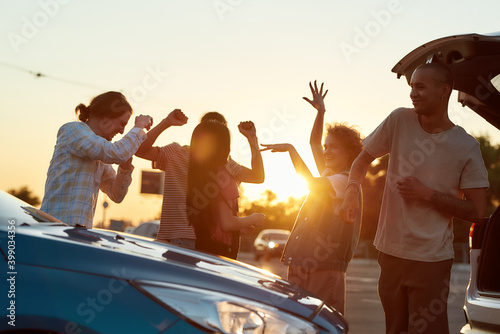 A group of young women and men dancing to music having a good time together outside on a parking site near their cars photo