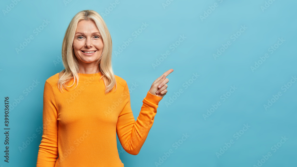 Φωτογραφία Waist up shot of blonde middle aged woman with glad expression shows awesome advertisement wears orange jumper isolated over blue background suggests to click link