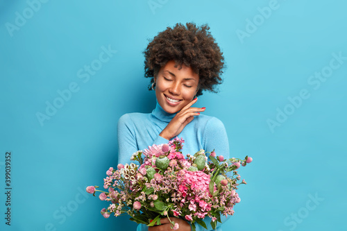 Portrait of gentle dark skinned woman touches jawline gently closes eyes and smiles pleasantly holds beeautiful bouquet of flowers has romantic mood isolated over blue background. Feminine concept
