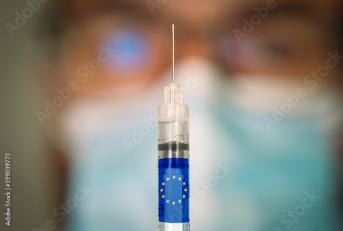 Close-up of hypodermic syringe with the European Union flag and a blurred doctor on the background. Selective focus. Concept of Covid vaccination campaign in Europe