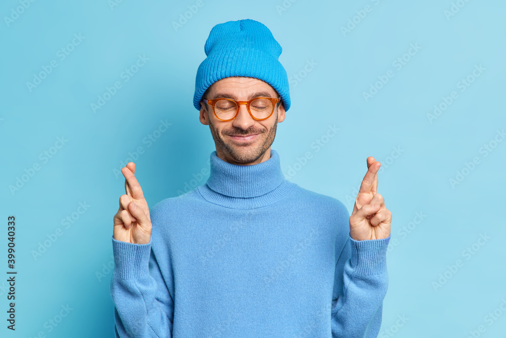 Portrait of glad European hipster man crosses fingers and smiles pleasantly believes in good luck wishes dreams come true wears stylish blue hat and poloneck stands indoor. Body language concept