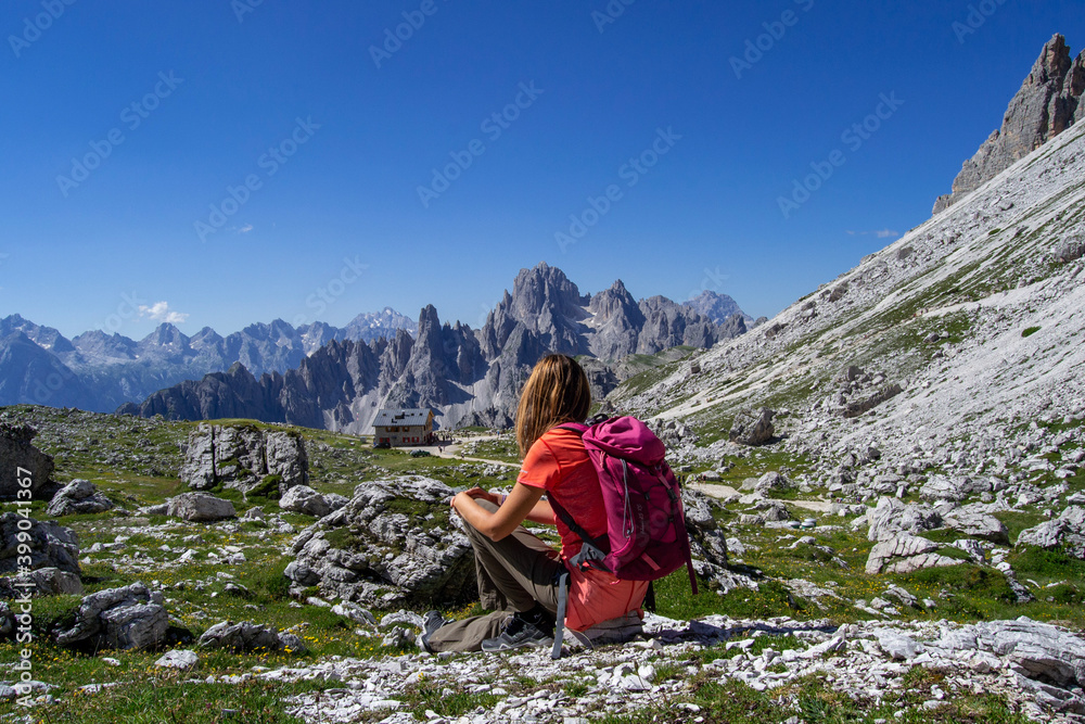 Young woman with backpack enjoying breathtaking view of Dolimites Alps mountain landscape sitting on a rock. Travel concept back view