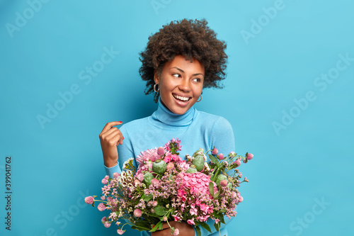 Portrait of dark skinned woman with curly hair looks gladfully away holds beautiful bouqet poses against blue background dressed casually. Millennial girl gets flowers from boyfriend on birthday