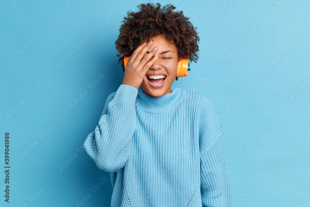 Happy joyful dark skinned woman with curly hair keeps hand on face smiles gladfully smiles broadly wears stereo headphones and casual jumper listens favorite music isolated on blue studio wall Happy joyful dark skinned woman with curly hair keeps hand on face smiles gladfully smiles broadly wears stereo headphones and casual jumper listens favorite music isolated on blue studio wall