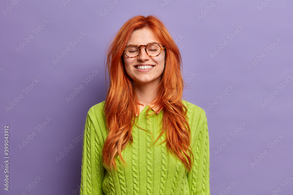 Relaxed beautiful redhead woman closes eyes and smiles broadly feels satisfied after hearing pleasant words wears green jumper and spectacles poses over vivid purple background. Happy emotions Relaxed beautiful redhead woman closes eyes and smiles broadly feels satisfied after hearing pleasant words wears green jumper and spectacles poses over vivid purple background. Happy emotions