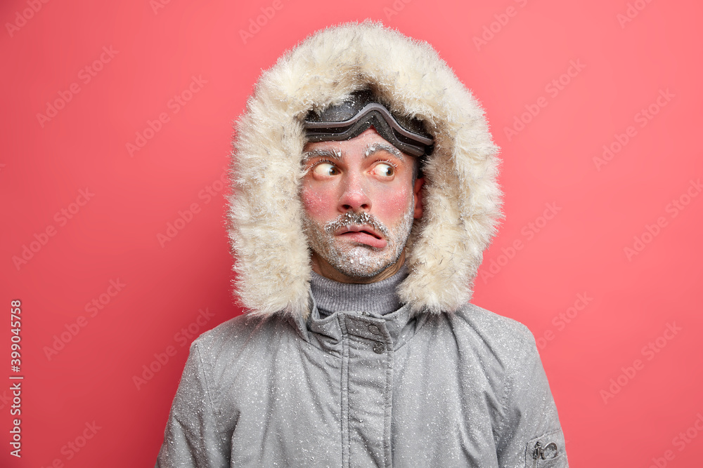 Headshot of emotive puzzled man purses lips and looks aside trembles from low temperature needs to warm wears grey jacket with fur hood poses indoor against pink background. Cold winter time Headshot of emotive puzzled man purses lips and looks aside trembles from low temperature needs to warm wears grey jacket with fur hood poses indoor against pink background. Cold winter time
