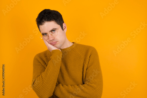 Very bored Young handsome Caucasian man wearing yellow sweater against orange wall holding hand on cheek while support it with another crossed hand, looking tired and sick. photo