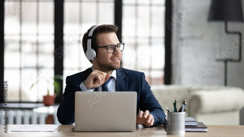 Dreamy businessman wearing headphones using laptop, sitting at desk in office, looking to aside, smiling employee executive dreaming, pondering future, listening to music after work done