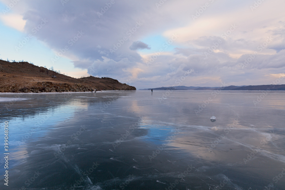 Beautiful winter landscape of frozen Lake Baikal with reflection of clouds in the mirror ice surface of the Kurkut Bay at sunset. Tourists walk on ice on a weekend. Natural background