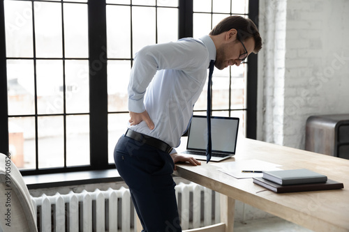 Unhealthy businessman wearing glasses touching lower back, suffering from backache, standing near desk in office, stressed employee feeling unhealthy and unwell after long hours sedentary work