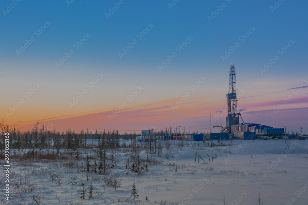 Winter landscape of a snowy forest tundra with a drilling rig for drilling an oil and gas well in the northern oil and gas field. Polar day sunset. Beautiful bright sky and clouds