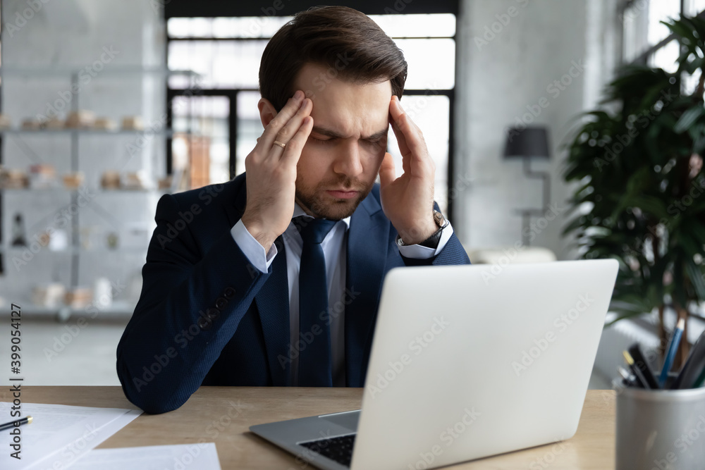 Tired stressed businessman touching massaging temples with closed eyes, sitting at desk with laptop, exhausted employee suffering from strong headache or migraine, relieving pain, blurred vision Tired stressed businessman touching massaging temples with closed eyes, sitting at desk with laptop, exhausted employee suffering from strong headache or migraine, relieving pain, blurred vision