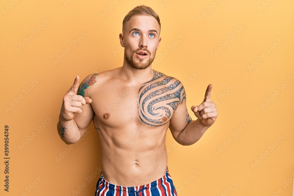 Fototapeta premium Young caucasian man wearing swimwear shirtless amazed and surprised looking up and pointing with fingers and raised arms.