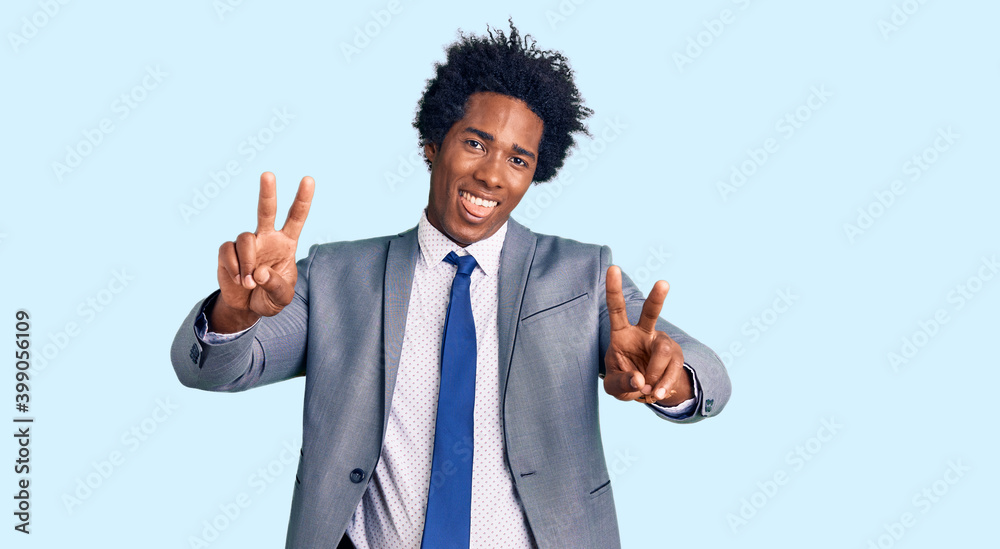 Handsome african american man with afro hair wearing business jacket smiling with tongue out showing fingers of both hands doing victory sign. number two.