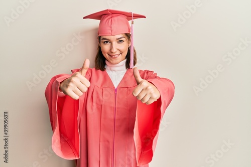 Young caucasian woman wearing graduation cap and ceremony robe approving doing positive gesture with hand, thumbs up smiling and happy for success. winner gesture.