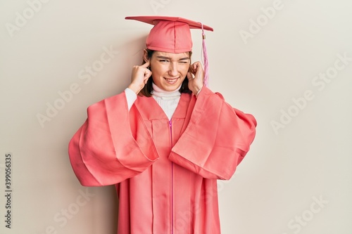 Young caucasian woman wearing graduation cap and ceremony robe covering ears with fingers with annoyed expression for the noise of loud music. deaf concept.