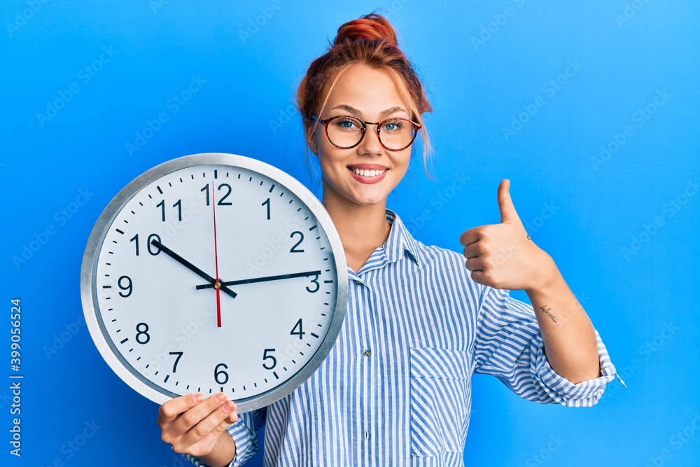 Young redhead woman holding big clock smiling happy and positive, thumb up doing excellent and approval sign Young redhead woman holding big clock smiling happy and positive, thumb up doing excellent and approval sign