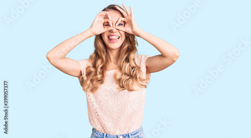 Young beautiful caucasian woman with blond hair wearing casual clothes doing ok gesture like binoculars sticking tongue out, eyes looking through fingers. crazy expression.