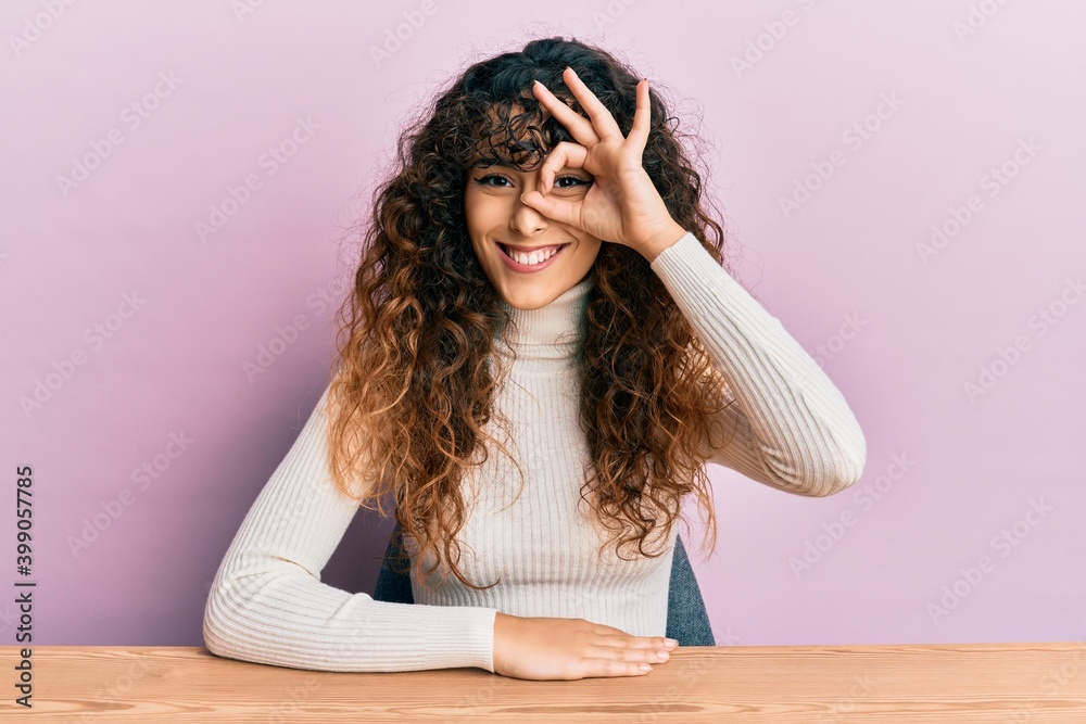 Young hispanic girl wearing casual clothes sitting on the table doing ok gesture with hand smiling, eye looking through fingers with happy face. Young hispanic girl wearing casual clothes sitting on the table doing ok gesture with hand smiling, eye looking through fingers with happy face.