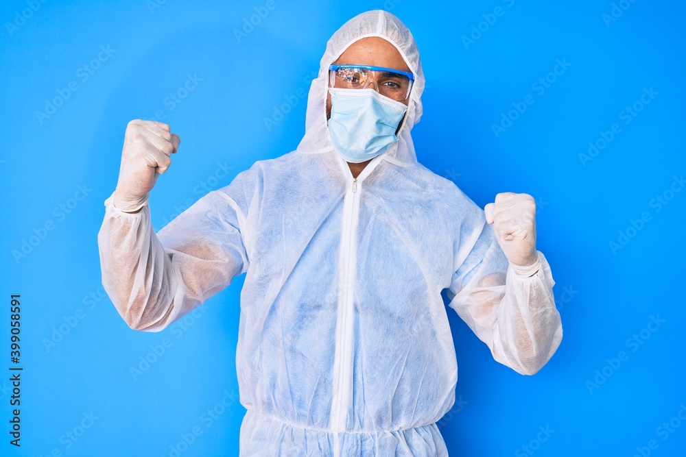 Young hispanic man wearing doctor coronavirus protection uniform screaming proud, celebrating victory and success very excited with raised arms Young hispanic man wearing doctor coronavirus protection uniform screaming proud, celebrating victory and success very excited with raised arms