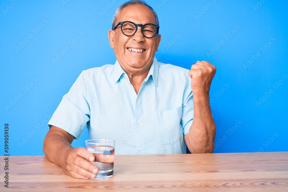 Senior handsome man with gray hair drinking glass of water sitting on the table screaming proud, celebrating victory and success very excited with raised arm Senior handsome man with gray hair drinking glass of water sitting on the table screaming proud, celebrating victory and success very excited with raised arm