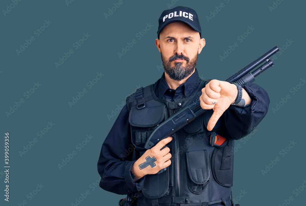 Young handsome man wearing police uniform holding shotgun with angry face, negative sign showing dislike with thumbs down, rejection concept Young handsome man wearing police uniform holding shotgun with angry face, negative sign showing dislike with thumbs down, rejection concept