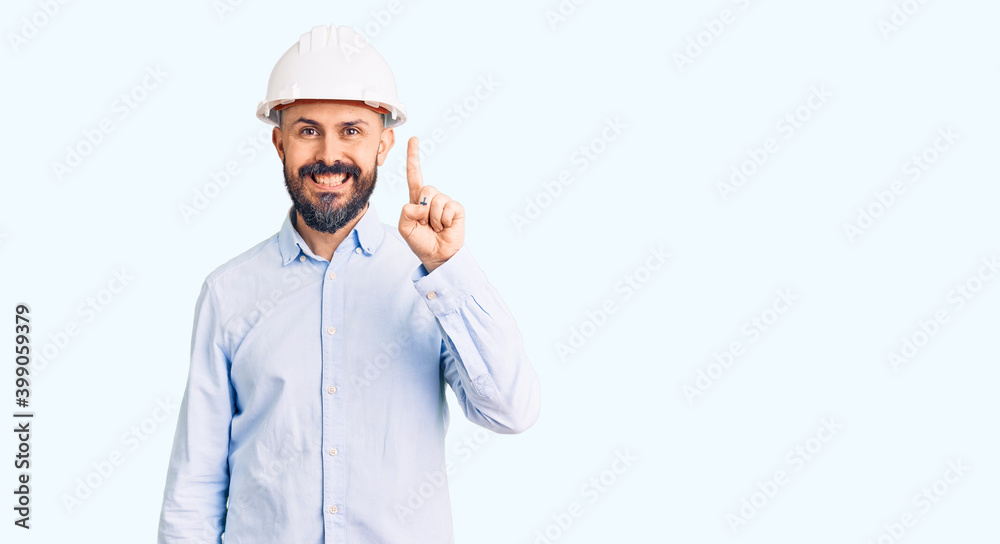 Young handsome man wearing architect hardhat showing and pointing up with finger number one while smiling confident and happy.