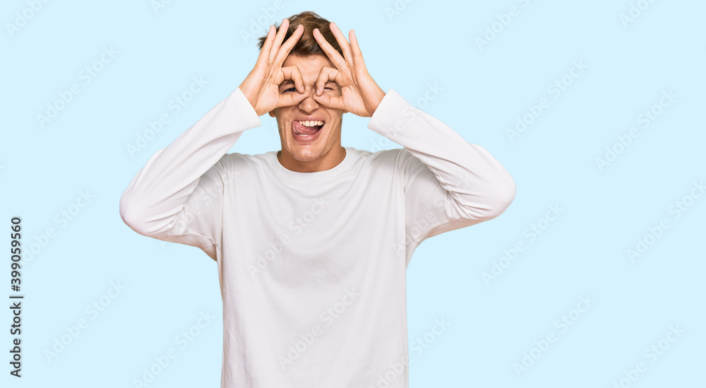 Handsome caucasian man wearing casual white sweater doing ok gesture like binoculars sticking tongue out, eyes looking through fingers. crazy expression. Handsome caucasian man wearing casual white sweater doing ok gesture like binoculars sticking tongue out, eyes looking through fingers. crazy expression.