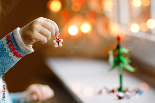Closeup of toddler girl by window and decorating small glass Christmas tree with tiny xmas toys. Child celebrate family traditional holiday. Close-up on hands with gingerbread man