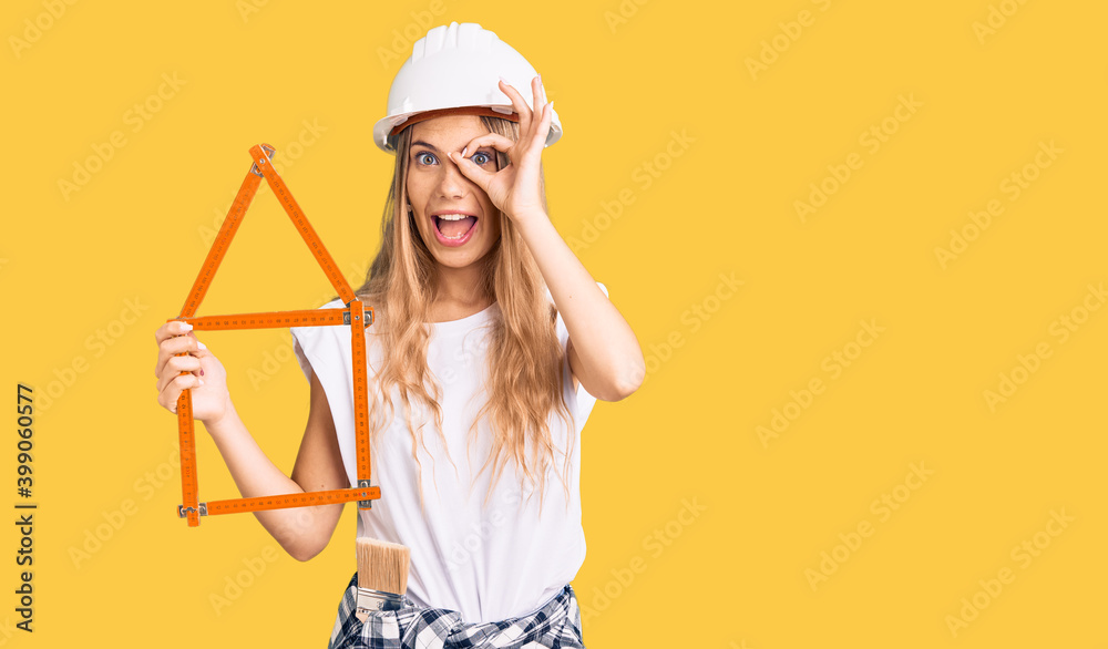 Beautiful caucasian woman with blonde hair wearing architect hardhat and holding tools smiling happy doing ok sign with hand on eye looking through fingers Beautiful caucasian woman with blonde hair wearing architect hardhat and holding tools smiling happy doing ok sign with hand on eye looking through fingers