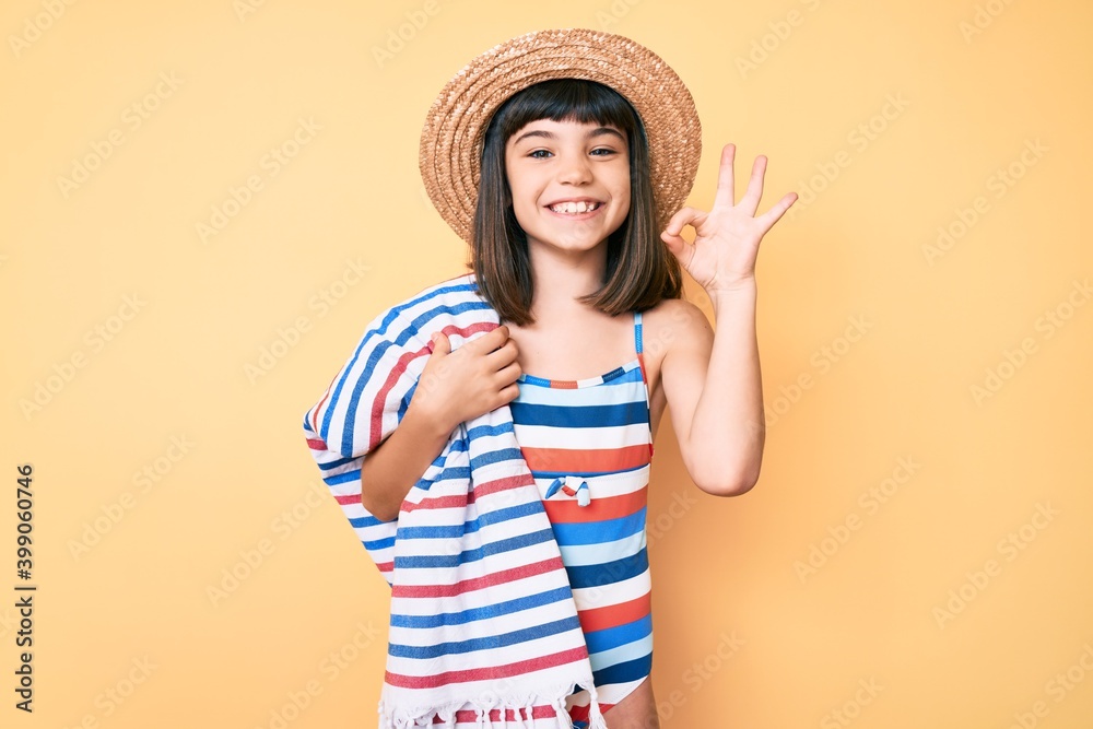 Young little girl with bang wearing swimsuit and summer hat holding towel doing ok sign with fingers, smiling friendly gesturing excellent symbol Young little girl with bang wearing swimsuit and summer hat holding towel doing ok sign with fingers, smiling friendly gesturing excellent symbol