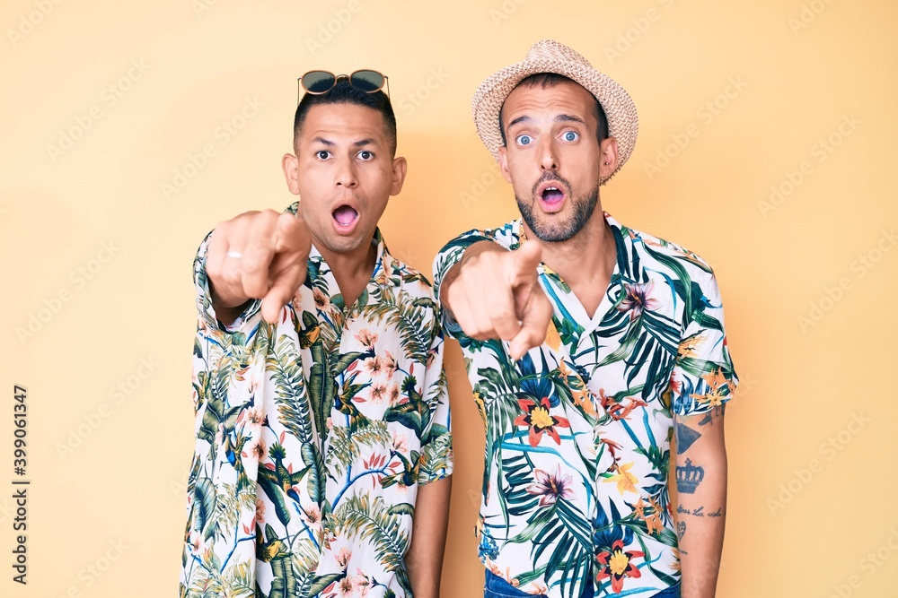 Young gay couple of two men wearing summer hat and hawaiian shirt pointing with finger surprised ahead, open mouth amazed expression, something on the front Young gay couple of two men wearing summer hat and hawaiian shirt pointing with finger surprised ahead, open mouth amazed expression, something on the front