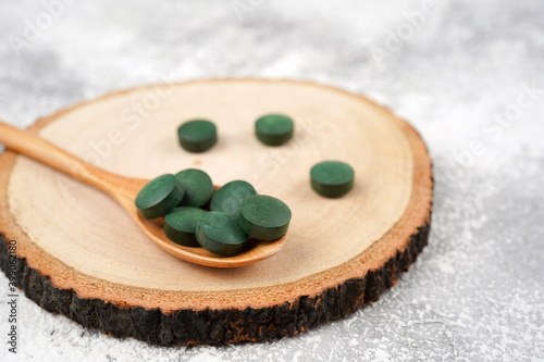 Spirulina tablets in wooden spoon on round stand, light concrete background. Nutrition, vitamin, immunity concept. Close-up, copy space