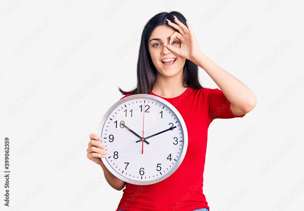 Young beautiful girl holding clock smiling happy doing ok sign with hand on eye looking through fingers Young beautiful girl holding clock smiling happy doing ok sign with hand on eye looking through fingers
