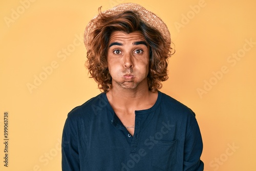 Vászonkép Young hispanic man wearing summer hat puffing cheeks with funny face