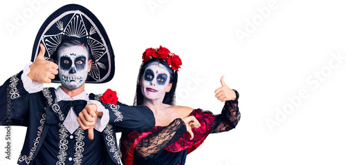 Young couple wearing mexican day of the dead costume over background doing thumbs up and down, disagreement and agreement expression. crazy conflict