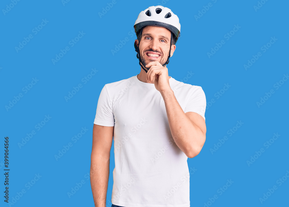Young handsome man wearing bike helmet looking confident at the camera with smile with crossed arms and hand raised on chin. thinking positive.