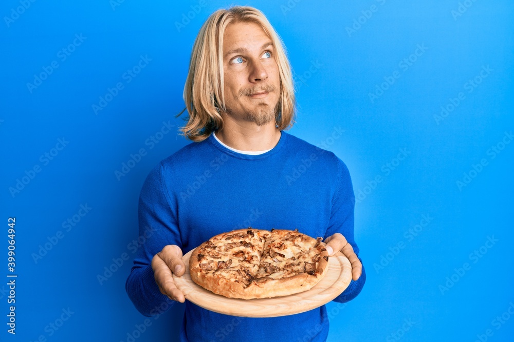 Caucasian young man with long hair holding italian pizza smiling looking to the side and staring away thinking. Caucasian young man with long hair holding italian pizza smiling looking to the side and staring away thinking.